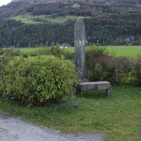 Concrete obelisk monument surrounded by hedges, with a bench inscribed in German, located at a rural crossroads near Oberurnen, Switzerland. Mountains and fields are visible in the background.