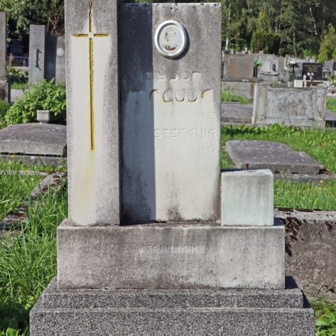 Gravestone of the Macuda family with a stele flanked by two rectangular blocks, the left one taller with a cross. An inscription and an oval photo above a palm branch are on the stele. The base has another inscription.