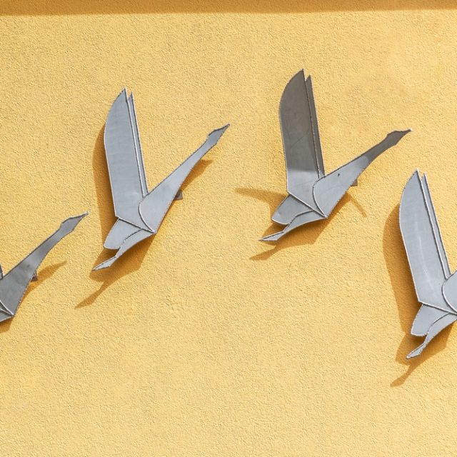 Aluminiowy relief 'Odlatujące łabędzie' Macieja Piotrowskiego na żółtej ścianie, przedstawiający cztery stylizowane łabędzie w locie, przypominające origami.