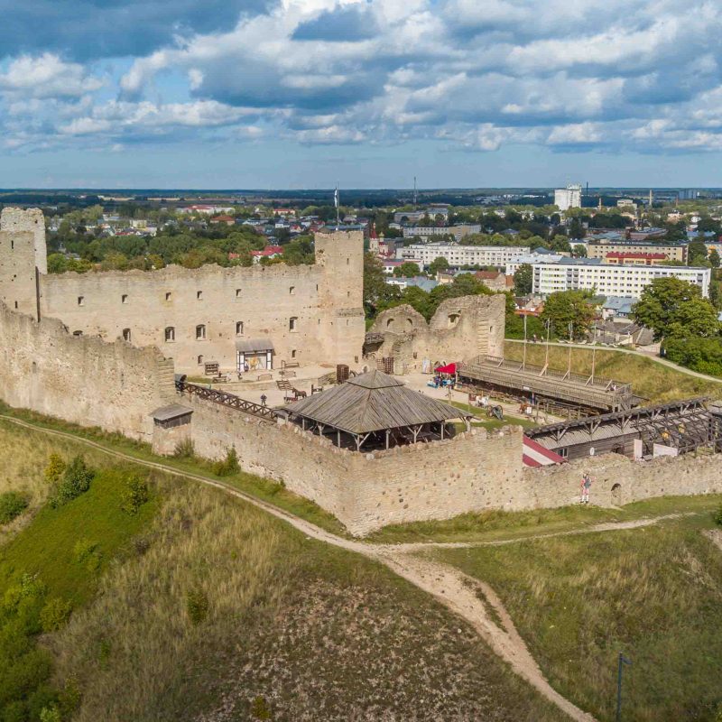 Vue aérienne du château de Rakvere, en Estonie, montrant ses murs médiévaux en pierre et la verdure environnante. À l'arrière-plan, on aperçoit la ville de Rakvere sous un ciel partiellement nuageux.