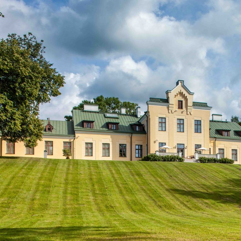 Le Musée de la guerre estonien à Viimsi, Estonie, situé dans un manoir historique avec une façade jaune et un toit vert, entouré d'une pelouse et d'arbres sous un ciel nuageux.