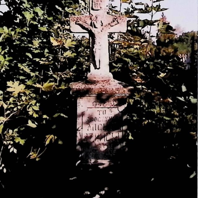 Gravestone of Michał Szelicki with a cross on a two-tiered pedestal. The cross features a sculpted figure of Christ and an inscription above. The pedestal has engraved inscriptions and is surrounded by foliage.