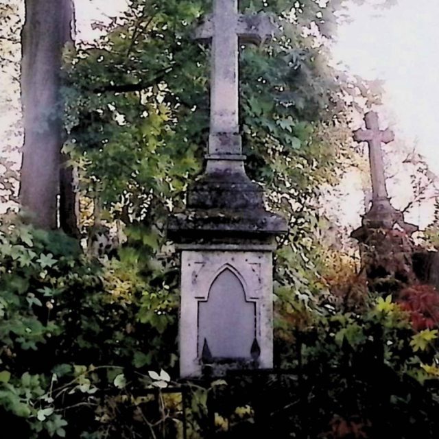 Gravestone with a cross on a two-tiered pedestal surrounded by foliage. The cross has zigzagged arms and sits on a small plinth. The pedestal features an arch-shaped panel with a sharp overhanging arch.