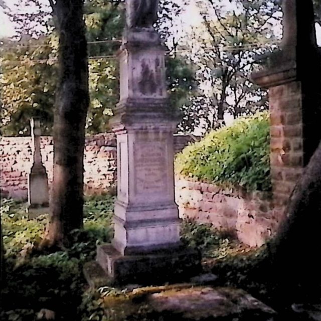 Gravestone of Aleksander Mrozowski with a two-tiered slab and a statue of the Virgin Mary with Child on a three-tiered pedestal. The statue is surrounded by trees and a stone wall.
