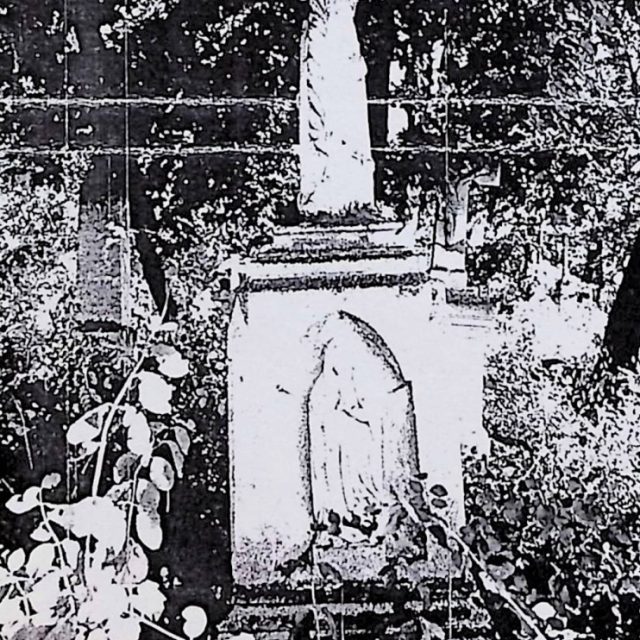 Tombe de Józef et Zuzanna Bojanowski avec une croix en forme de tronc d'arbre sur un piédestal à deux niveaux. La croix présente une feuille de palmier, des branches de rose et des fleurs. Une niche avec un relief de Notre-Dame du Rosaire est visible.