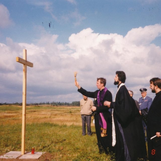 A group of people, including clergy in robes, gather around a wooden cross during a ceremony on a grassy field. A candle is placed at the base of the cross.