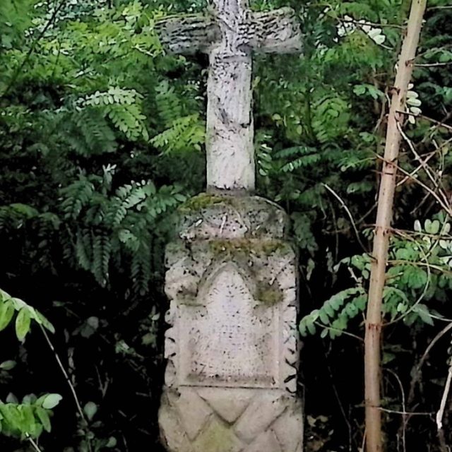 Gravestone with a cross styled as a tree trunk on a pedestal resembling rocks, surrounded by foliage. The cross is entwined with vine branches, and an inscription is carved on the front panel.