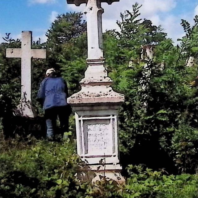 Gravestone of Bazyli Monasterski with a cross on a two-tier pedestal, featuring an octagonal cross-section and INRI inscription. Surrounded by greenery, a person stands nearby.