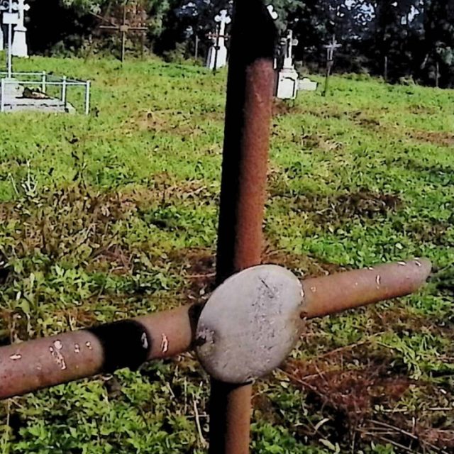 Iron cross on a pedestal marking the grave of Szczepan Korczyński. The cross is tubular with an oval metal plaque at the intersection of the arms. The grave is overturned, with parts lying separately.