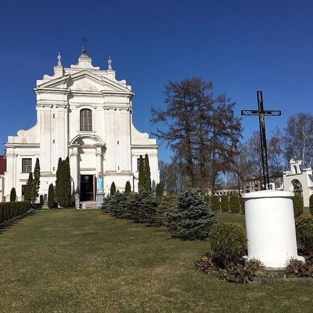 Костел святого Людовика в Краславі, біла барокова будівля з симетричним фасадом, оточена деревами та газоном. На передньому плані чорний хрест на білому постаменті.