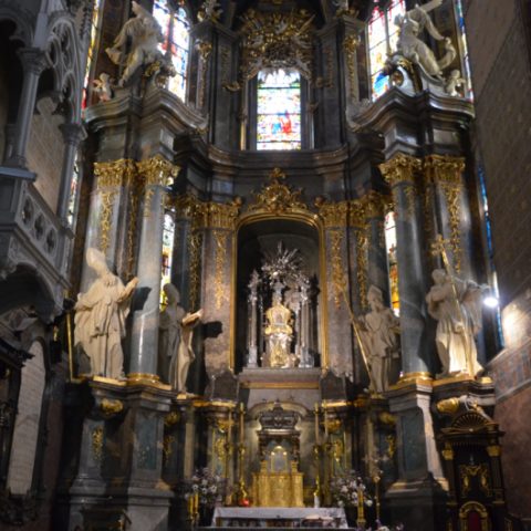 The high altar of the Latin Cathedral in Lviv, featuring ornate columns, statues of the Church Fathers and a central niche with a silver halo. The stained-glass windows bathe the space in light.
