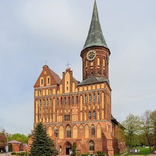 La cathédrale gothique de la Bienheureuse Vierge Marie et Saint-Adalbert à Kaliningrad, avec une haute flèche dotée d'une horloge, une façade en briques rouges et des fenêtres en arc. Des arbres et un chemin au premier plan.