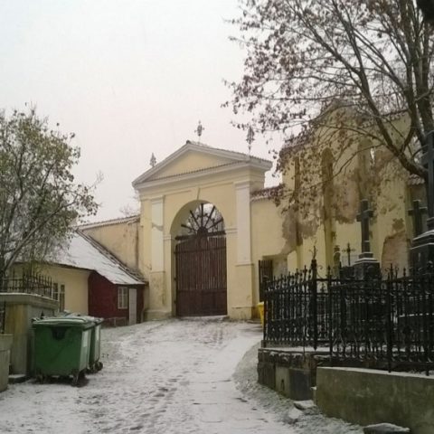 The entrance gate to the Bernardine Cemetery in Vilnius, surrounded by snow-covered ground and trees. Iron crosses and a fence can be seen on the right.