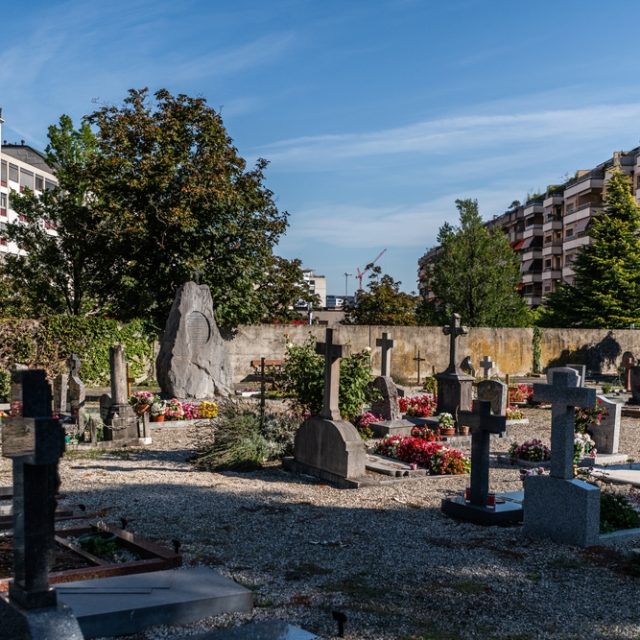 Nagrobek generała Józefa Hauke-Bosaka na cmentarzu w Carouge, otoczony innymi nagrobkami i kwiatami. Kamień jest duży, w kształcie piramidy z żelaznym krzyżem na szczycie. W tle widoczne są budynki mieszkalne.