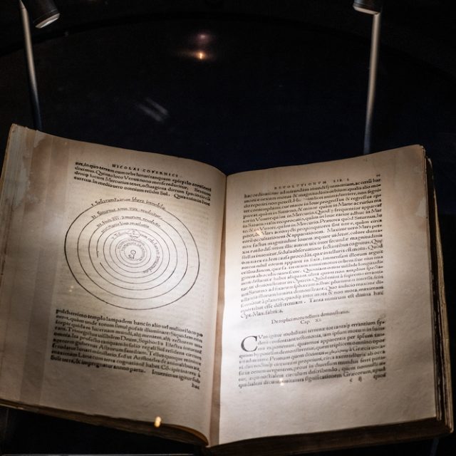 Livre ouvert, dont la page visible est tirée de *De revolutionibus orbium coelestium* de Nicolas Copernic, comportant un diagramme héliocentrique et un texte en latin, éclairé.