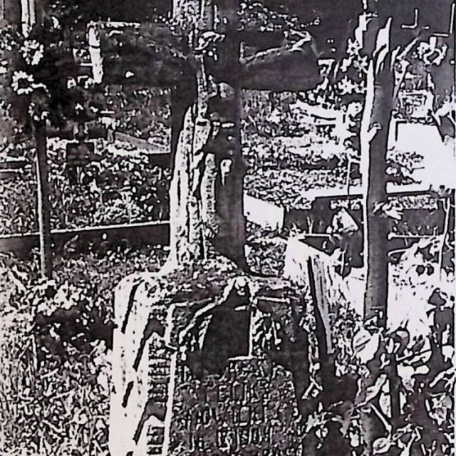 Tombe marquée par une croix sur un piédestal, stylisée comme des poutres d'arbre avec une figure sculptée du Christ à l'intersection. Au-dessus, une grenade fissurée. En dessous, une plaque en forme de bouclier avec une inscription et des branches de chêne.