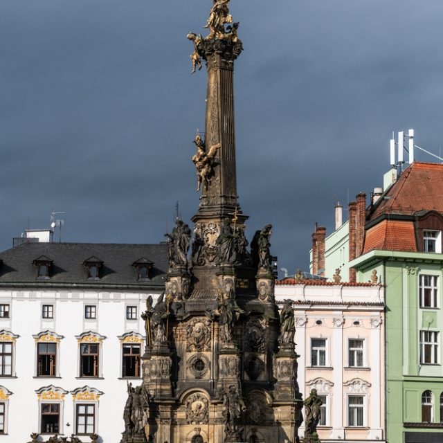The Holy Trinity Column in Olomouc, a 32-meter baroque monument with sculptures of saints, stands on the Upper Square. It is surrounded by historic buildings under a cloudy sky.