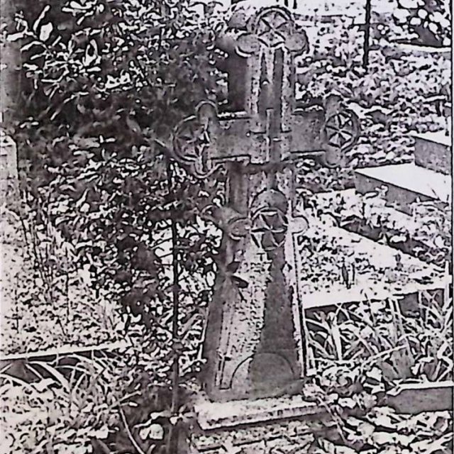 Gravestone with a cross on a pedestal, featuring trefoil-ended arms and a rosette. The cross has a carved groove and a smaller cross at the intersection. Below is a rosette and an inscription.