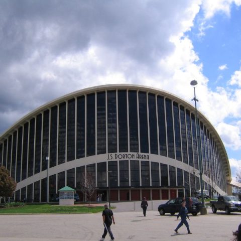 Dorton Arena à Raleigh avec ses arcs paraboliques distinctifs et sa façade en verre sous un ciel nuageux. Des voitures et des personnes sont visibles au premier plan.