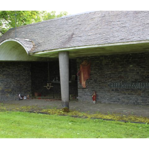 The Notre-Dame-de-la-Foi Chapel in Bertrix, Belgium, has a stone façade featuring terracotta sculptures of angels and the Virgin Mary. A metal grille and a single column support the structure. It is set in a grassy setting.