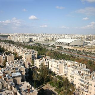 Aerial view of Aleppo with a prominent sports hall and swimming pool designed by Polish architect Wojciech Zabłocki. The large, dome-shaped structure stands out amidst the urban landscape.