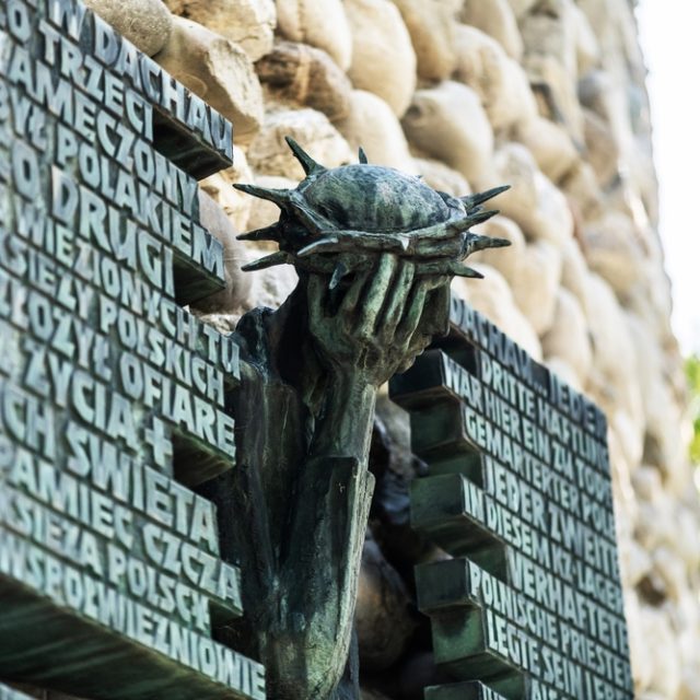 Sculpture en bronze du Christ souffrant avec une couronne d'épines, tenant sa tête dans ses mains. Inscription en polonais et en allemand sur des plaques à côté de lui. Située sur le mur arrière de la chapelle du camp de concentration de Dachau.