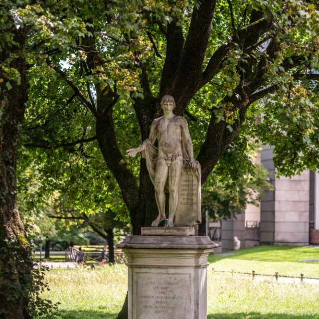 Statue d'un jeune homme connu sous le nom de 'Niewinny' à Munich, debout sur un piédestal dans une zone verte avec des arbres. La figure est nue et s'appuie sur une tablette en pierre avec une inscription.