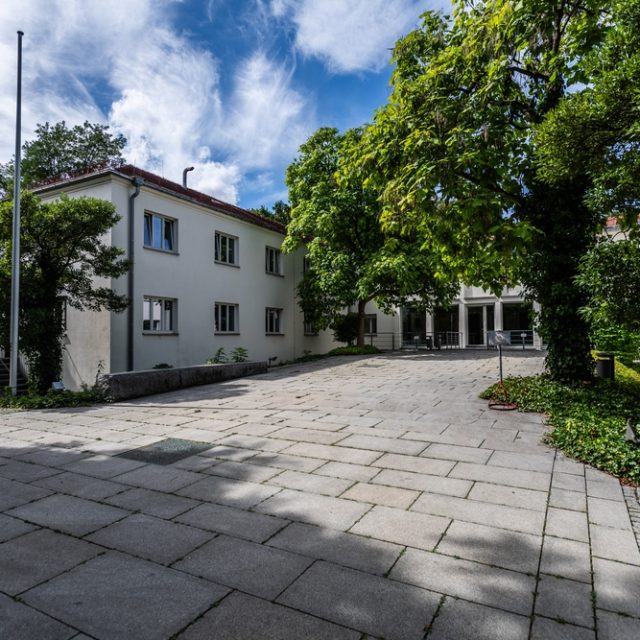 L'ancien siège de Radio Free Europe à Munich, un bâtiment moderniste entouré d'arbres, avec une cour pavée et des mâts de drapeau.
