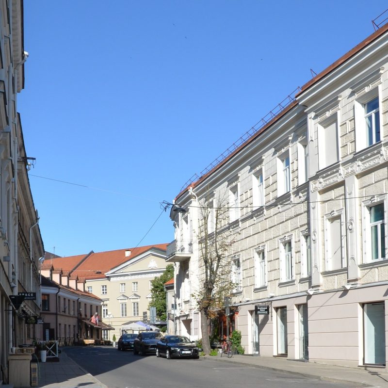 Street view in Vilnius with the Ogiński Palace in the background. The street is lined with historic buildings and parked cars. The palace features a light facade with a triangular pediment.