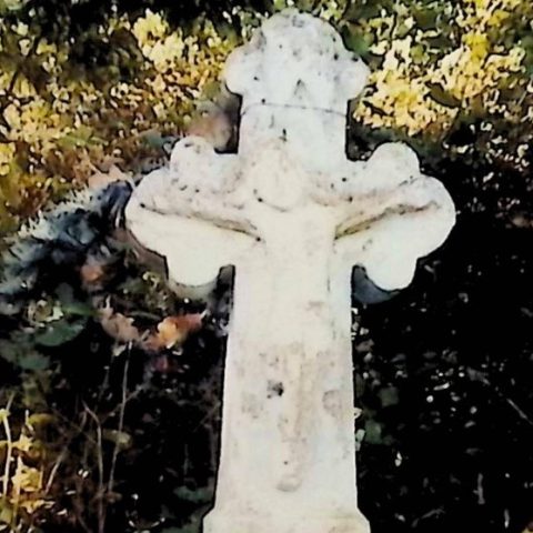 Gravestone with a cross on a pedestal, featuring a trefoil-ended cross with a relief of Christ at the intersection. Surrounded by foliage.