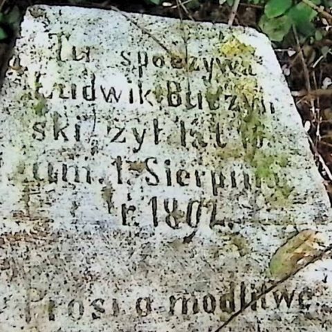 Tombstone of Ludwik Buczyński with engraved inscription, partially covered by moss. The stone is weathered and surrounded by vegetation.
