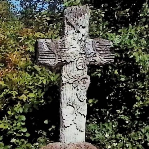 Gravestone with a cross styled as a tree trunk on a rock-like pedestal. The cross is decorated with carved oak branches and acorns. The base has beveled top edges. A scroll-shaped plaque with an inscription is on the front.