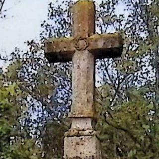 Gravestone with a cross on a two-tiered pedestal surrounded by trees. The cross has octagonal arms and an inscription on the front panel.