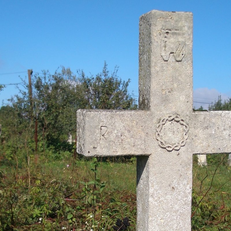 Kamienny krzyż na cokole z reliefem korony cierniowej na przecięciu ramion, literami INRI i czaszką z piszczelami poniżej inskrypcji. Otoczony zielenią.