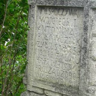 Pierre tombale d'Antonina et Wojciech Połowczuk avec une inscription dans un panneau encadré. Une croix cassée repose à côté de la pierre, initialement stylisée comme un tronc d'arbre sur un socle rocheux.