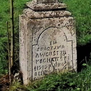 Nagrobek Augustyna Kochańskiego z krzyżem na cokole. Krzyż ma ramiona zakończone łukiem kotarowym. Na froncie wyryta inskrypcja w łukowej ramie.