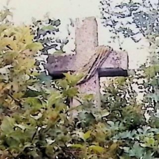Kamienny krzyż z ramionami zakończonymi nasadkami, szerszy u dołu, na dwustopniowym cokole otoczony zielenią. Przez ramiona przewieszona wstęga reliefowa.