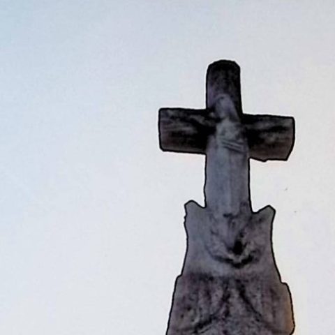 Gravestone with a cross styled as a tree trunk, featuring Christ at the intersection of arms. A wreath of leaves below. The pedestal resembles a stone pile with an inscription on a scroll.