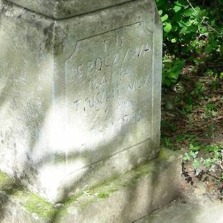 Nagrobek z krzyżem na cokole, z reliefem Chrystusa i inskrypcją. Cokół ma ścięte i profilowane krawędzie, otoczony zielenią.