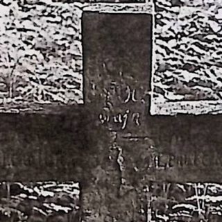 Grób oznaczony prostym krzyżem z wyrytą inskrypcją, otoczony trawą.