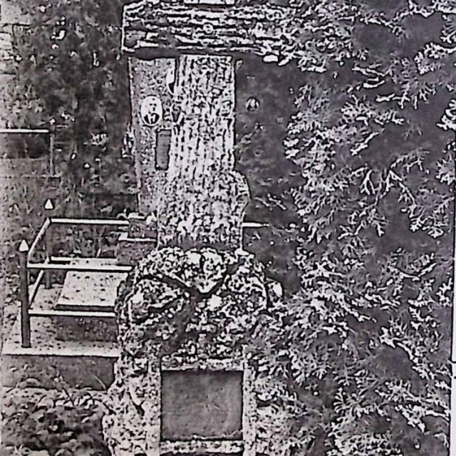 Gravestone of Daria Krajewska with a cross resembling a tree trunk on a stone pile base. A red sandstone panel with an engraved inscription and a raised cross is on the front. Six holes form a cross shape on the plate.