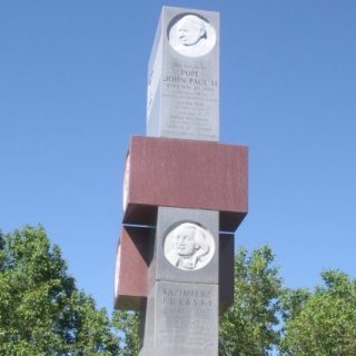 Pomnik w Parku Pułaskiego w Denver z szarego i różowego granitu, z medalionami wybitnych Polaków, w tym Jana Pawła II i Kazimierza Pułaskiego. W tle drzewa i niebieskie niebo.