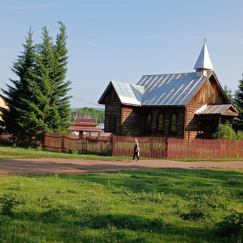 Wooden Church of St. Stanislaus Bishop and Martyr in Wierszyna, surrounded by a wooden fence and trees, with a person walking nearby.