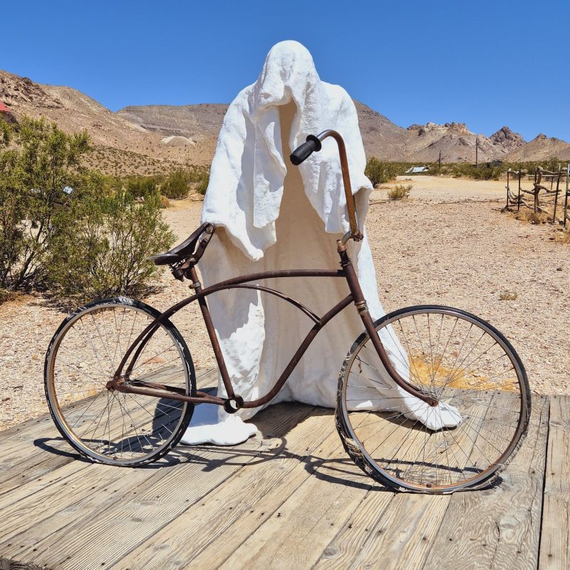 Rzeźba 'Ghost Rider' Charlesa Alberta Szukalskiego w Rhyolite, Nevada, przedstawiająca postać w białym całunie stojącą za zardzewiałym rowerem na drewnianej platformie.