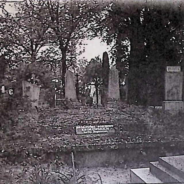 Tombeau rectangulaire de la famille Krokowski, plâtré et couvert d'une dalle rectangulaire plus large. Une plaque secondaire avec une inscription est à l'avant. Entouré d'arbres et d'autres pierres tombales.