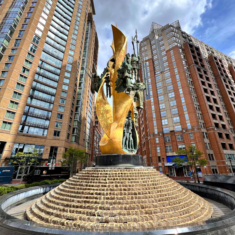 The National Katyn Memorial in Baltimore, known as the 'Flame of Freedom,' features a bronze sculpture with golden flames and figures of Polish officers. It stands on a stepped base, surrounded by tall buildings.