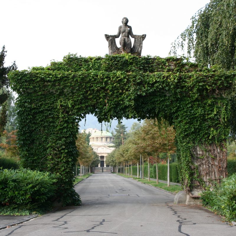 Le portail d'entrée recouvert de lierre du cimetière de Silhfeld à Zurich, surmonté d'une sculpture et bordé d'un allée menant au bâtiment de style classique visible à l'arrière-plan.
