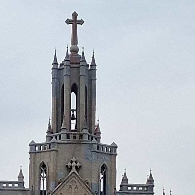 Le sommet de l'église polonaise (cathédrale du Sacré-Cœur de Jésus) à Tachkent, avec une haute flèche ornée d'une croix et de pinacles décoratifs sous un ciel nuageux.