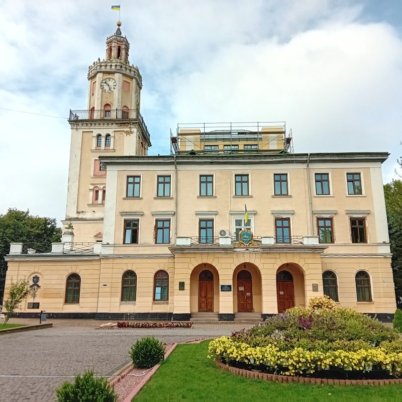Hôtel de ville de Sambor avec une haute tour d'horloge et une façade beige, entouré de verdure et d'un parterre de fleurs au premier plan.