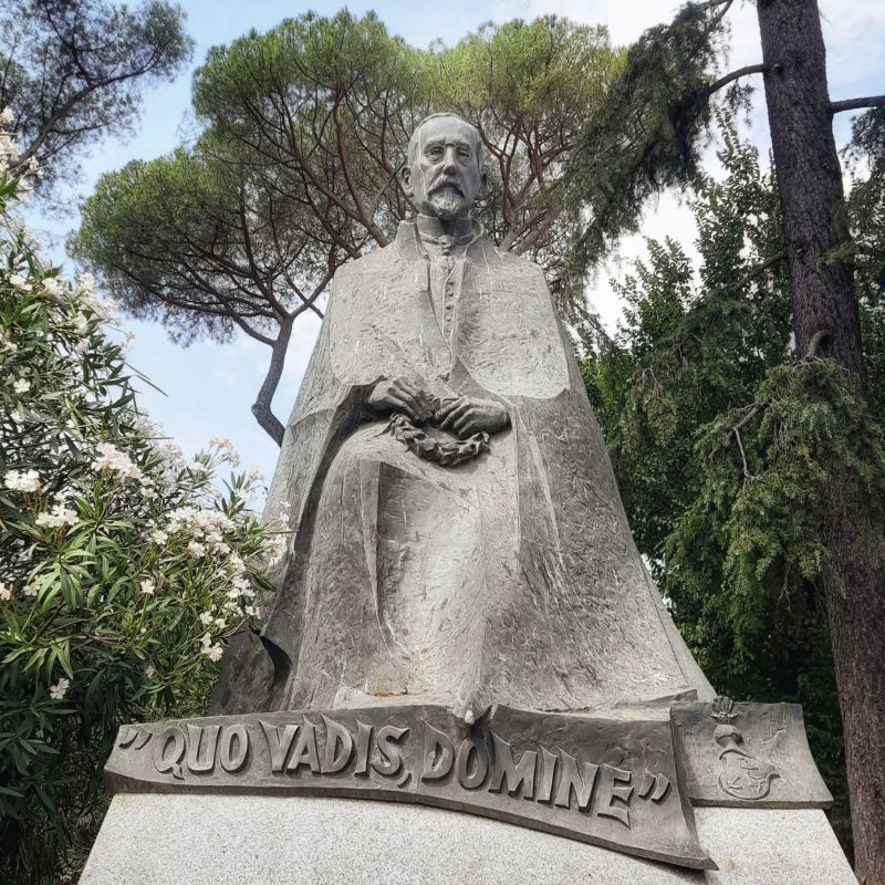 Pomnik Henryka Sienkiewicza trzymającego wieniec laurowy, otoczony drzewami i kwiatami w Ogrodach Villi Borghese.